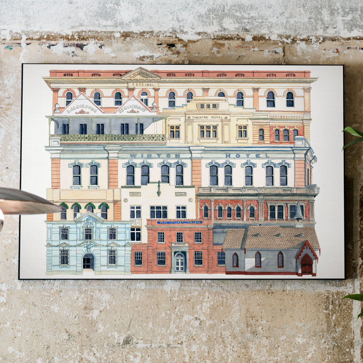 A framed art print depicting a row of colorful vintage buildings, displayed on a textured concrete wall next to a hanging lamp and green plant leaves.